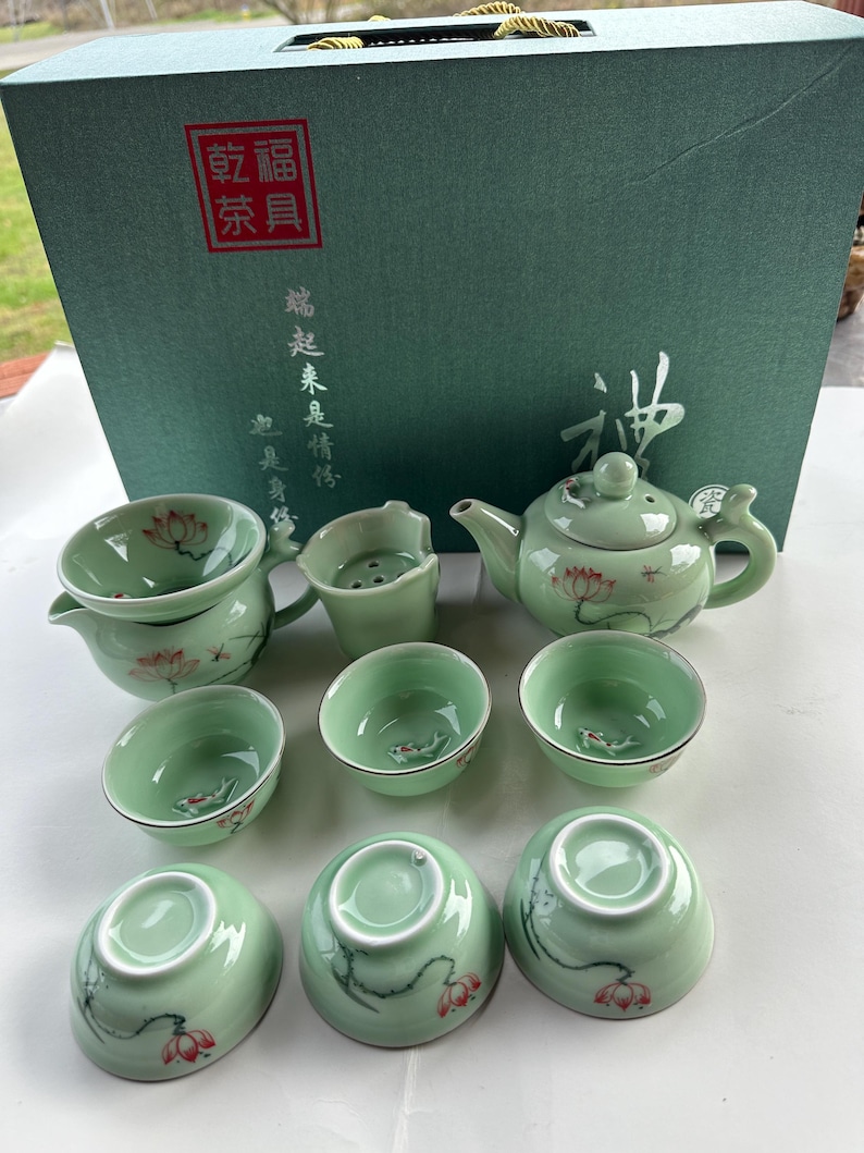 May include: A green tea set with a teapot, a strainer, and six cups. The set is decorated with a lotus flower design. The box is green and has Chinese characters on it.