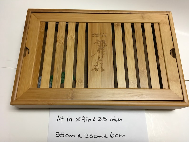 May include: Rectangular bamboo tea tray, 14 inches long, 9 inches wide, and 2.5 inches high. The tray features a slatted design with a central panel etched with a bamboo plant and Chinese characters. The tray has rounded corners and handles.