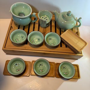 May include: A complete tea set in a light green glaze, including a teapot, strainer, and several small cups. Each cup features a koi fish design. The set is arranged on a bamboo tray, with additional bamboo coasters for the cups.