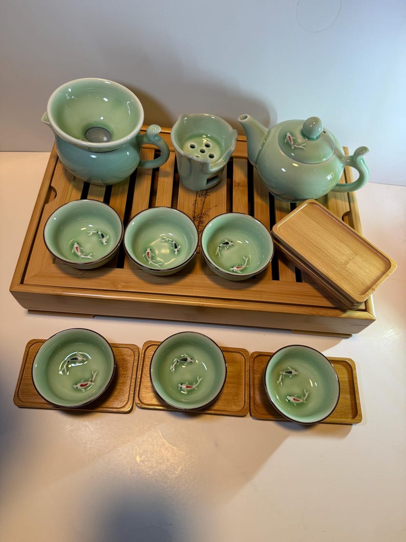 May include: A complete tea set in a light green glaze, including a teapot, strainer, and several small cups. Each cup features a koi fish design. The set is arranged on a bamboo tray, with additional bamboo coasters for the cups.