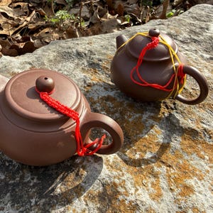 May include: Two brown ceramic teapots with rounded bodies and curved handles. Each teapot has a lid with a small knob and is adorned with a decorative red or yellow cord. The teapots are displayed on a large, textured rock.