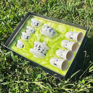 May include: A white ceramic tea set with black calligraphy in a gift box. The set includes a teapot, a teacup with a saucer, and six small teacups. The box is lined with a bright green fabric, and the set is displayed on a bed of green grass.