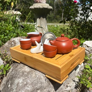 May include: A complete tea set on a bamboo tray. The set includes a reddish-brown teapot, two small teacups, a creamer, and a metal strainer. A white ceramic sculpture is also included. The tea set is displayed outdoors on a rock.