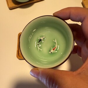 May include: A light green ceramic teacup with a brown rim, featuring two small koi fish figurines inside. The teacup is held by a hand and rests on a small wooden coaster. The teacup has a glossy finish.
