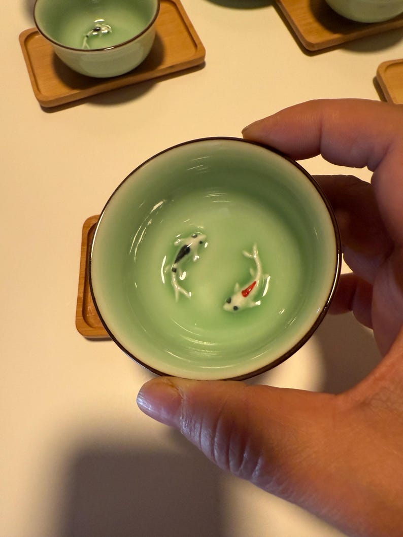May include: A light green ceramic teacup with a brown rim, featuring two small koi fish figurines inside. The teacup is held by a hand and rests on a small wooden coaster. The teacup has a glossy finish.