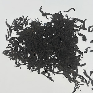May include: Close-up of loose-leaf tea. The tea leaves are dark brown and appear dried. The leaves are scattered across a white background, with some leaves appearing to be curled.
