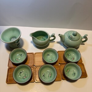 May include: A complete tea set in a soft green hue, featuring a teapot, a pitcher, a strainer, and six small cups. Each cup and the teapot lid are adorned with a koi fish design. The cups rest on individual wooden coasters.