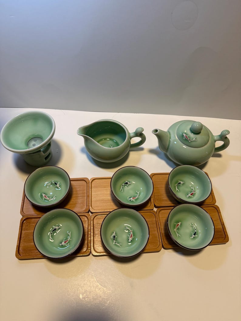 May include: A complete tea set in a soft green hue, featuring a teapot, a pitcher, a strainer, and six small cups. Each cup and the teapot lid are adorned with a koi fish design. The cups rest on individual wooden coasters.