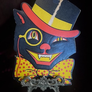 May include: A vintage Halloween decoration featuring a black cat with a top hat, monocle, and bow tie. The cat's face is smiling with sharp teeth. The bow tie is yellow with red polka dots. The decoration is displayed on a decorative metal stand.