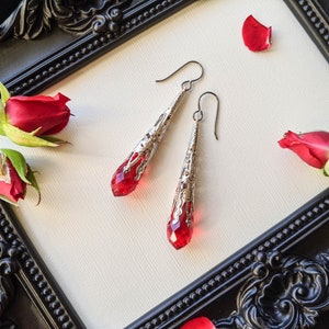 May include: A pair of silver dangle earrings with red crystal teardrop accents. The earrings are displayed on a white background with red roses.