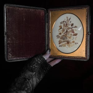 May include: An antique, oval-shaped picture frame with a gold-colored frame and a velvet-lined case. The frame contains a pressed flower arrangement with dried leaves and flowers.