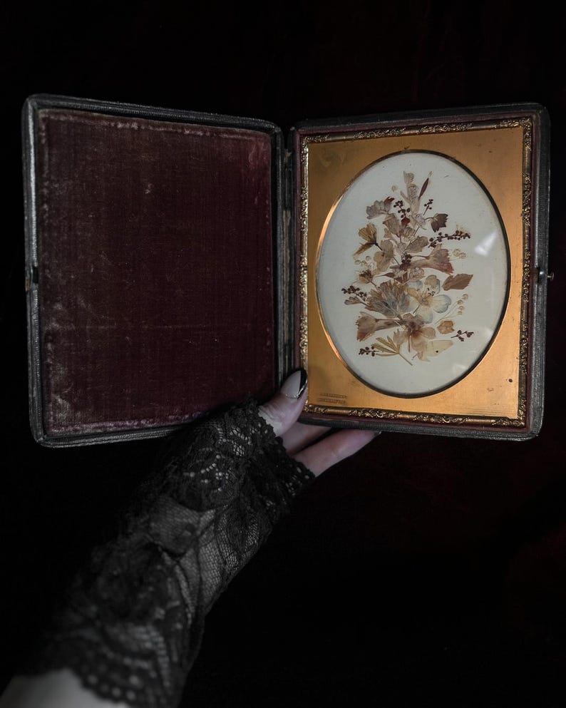 May include: An antique, oval-shaped picture frame with a gold-colored frame and a velvet-lined case. The frame contains a pressed flower arrangement with dried leaves and flowers.