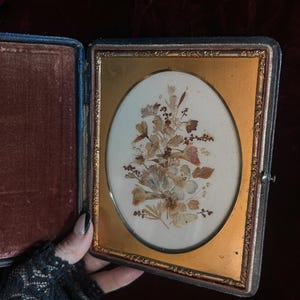 May include: A vintage oval-shaped pressed flower picture frame with a gold-colored frame and a dark brown velvet-lined case.