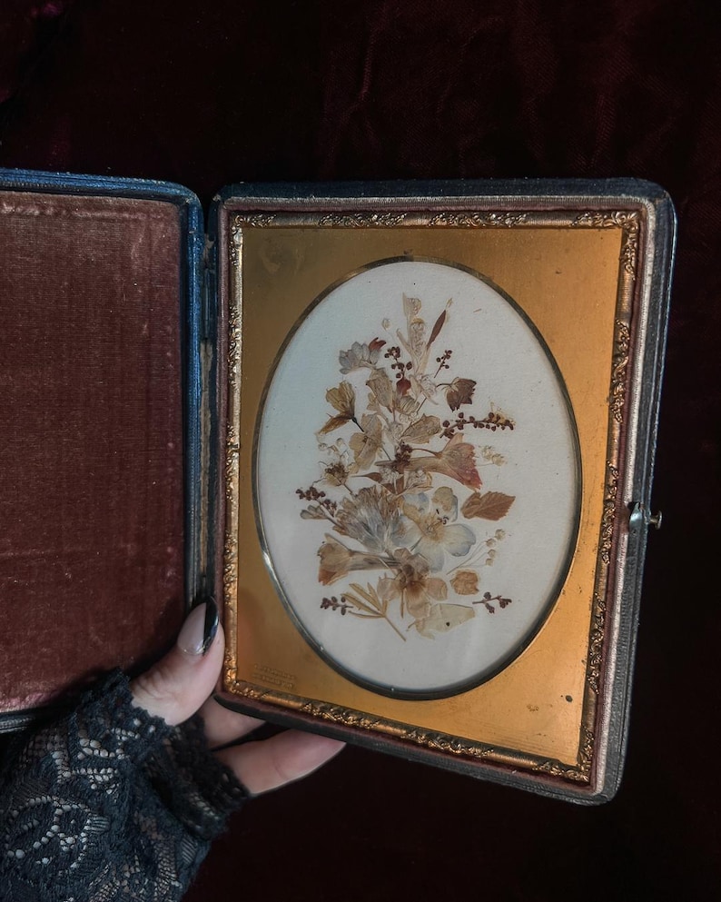 May include: A vintage oval-shaped pressed flower picture frame with a gold-colored frame and a dark brown velvet-lined case.