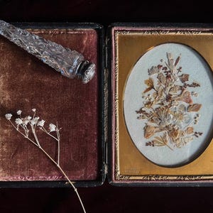 May include: A vintage, framed pressed flower art piece with a gold frame and a velvet-lined box with a glass bottle stopper. The pressed flowers are arranged in a bouquet shape and are a light brown color. The bottle stopper is clear glass with a silver top.