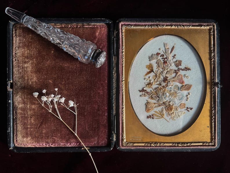 May include: A vintage, framed pressed flower art piece with a gold frame and a velvet-lined box with a glass bottle stopper. The pressed flowers are arranged in a bouquet shape and are a light brown color. The bottle stopper is clear glass with a silver top.