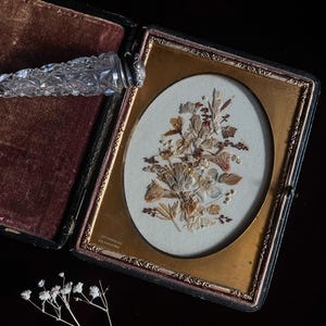 May include: An antique, black velvet-lined, hinged photo frame with a gold-toned oval frame. The frame contains a pressed flower arrangement with dried leaves and flowers. The frame is open and the inscription "HAMILTON, ONT" is visible on the bottom.