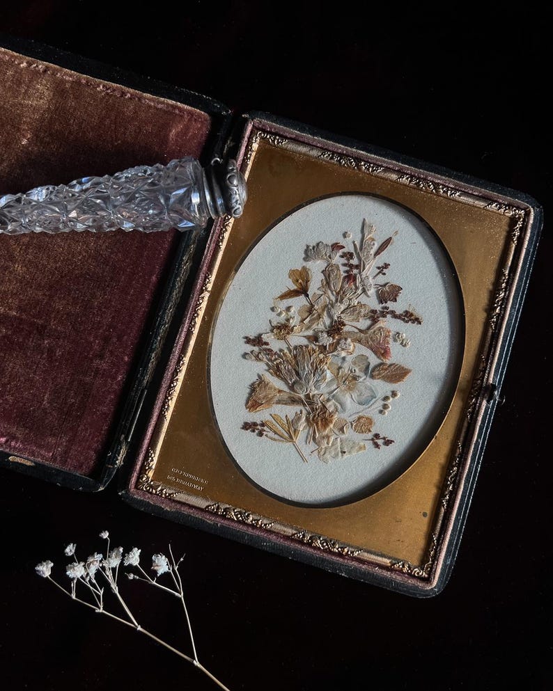 May include: An antique, black velvet-lined, hinged photo frame with a gold-toned oval frame. The frame contains a pressed flower arrangement with dried leaves and flowers. The frame is open and the inscription "HAMILTON, ONT" is visible on the bottom.