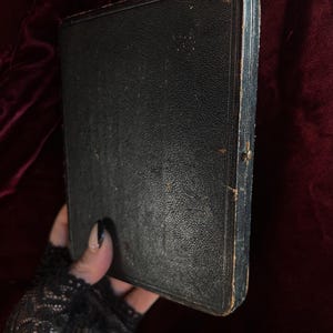 May include: A black leather-bound book with a worn, aged appearance. The book is closed and has a simple, rectangular shape.