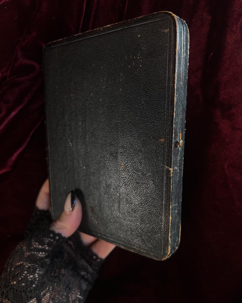May include: A black leather-bound book with a worn, aged appearance. The book is closed and has a simple, rectangular shape.