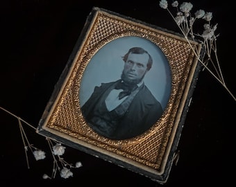 May include: A black and gold framed portrait of a man with a beard and mustache. The man is wearing a dark suit and a white shirt with a patterned vest. The portrait is in an oval frame.