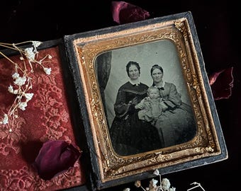 May include: Antique framed photograph featuring two women and a baby. The portrait is in a gold-toned frame with ornate detailing. The photograph is set against a red velvet background with dried flowers and rose petals.