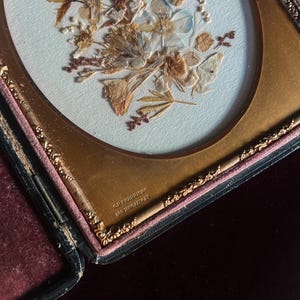 May include: A framed pressed flower arrangement in a gold oval frame with a black velvet backing. The frame has the inscription "G.F. HERRING 152 BROADWAY" on the bottom.