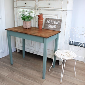 May include: A light blue wooden table with a brown top. The table has four legs and is standing on a wooden floor. There is a white sign that says "Potting Shed" on the table. There are also some terracotta pots on the table.