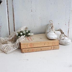 Könnte beinhalten: Zwei antike Bücher mit französischen Titeln, "Le Mobilier De Satin" und "Le Soulier De Satin", übereinander gestapelt. Ein Paar weiße Babyschuhe liegen auf den Büchern.