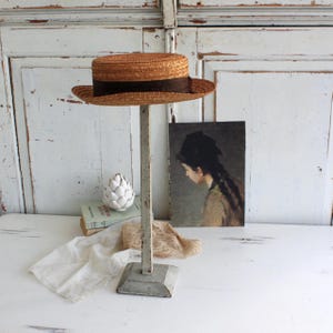 May include: A straw boater hat with a dark brown band sits atop a distressed, off-white metal hat stand. The hat stand has a square base and a vertical pole. A vintage painting and decorative objects are in the background.