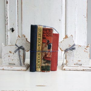 Puede incluir: Un par de sujetalibros blancos pintados con forma de perros, sosteniendo una pila de cuatro libros. Los libros se titulan "Story Book for Schoolboys", "The Big Budget for Boys" y "Blackie".