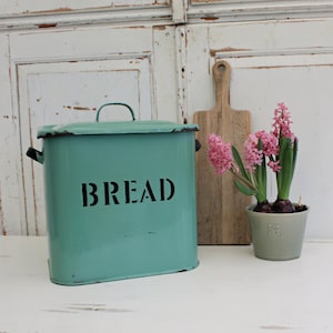 May include: A teal bread box with the word "BREAD" in black lettering. The bread box has a handle on top and two side handles. A wooden cutting board and a potted plant with pink flowers are next to the bread box.