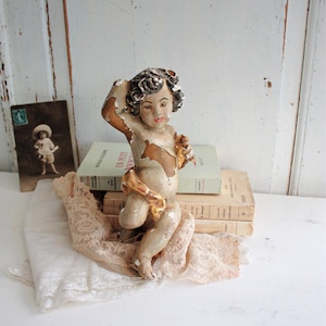Puede incluir: Una estatuilla de querubín vintage con pintura descascarillada, cuerpo crema, detalles marrones y una faja dorada. El querubín está posando con un brazo levantado. Descansa sobre un paño de encaje, con libros y una postal vintage al fondo.