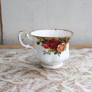 Pode incluir: Uma chávena de chá branca com uma borda dourada e um padrão floral com rosas vermelhas, amarelas e cor-de-rosa. A chávena tem uma pega delicada e uma base festoneada.