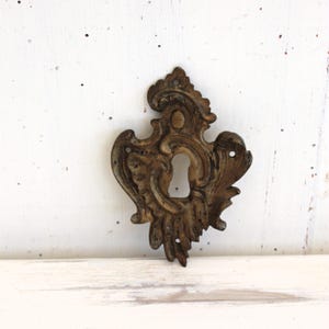May include: Ornate, antique-style keyhole cover in a bronze finish. The decorative piece features a central keyhole surrounded by swirling, floral-inspired designs. The metalwork has a weathered appearance, suggesting age and vintage charm.