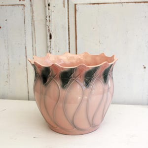 Pode incluir: Um vaso de cerâmica rosa com uma borda ondulada e um padrão decorativo. O vaso apresenta uma base rosa claro com um design de folhas rosa escuro e verde ao redor da parte superior. O interior é rosa claro sólido.