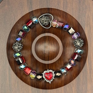 May include: A beaded bracelet featuring faceted red, iridescent, and dark metallic beads. The design includes a central silver-toned charm with a red heart-shaped stone and a silver-toned heart-shaped clasp at the top, creating a circular arrangement.