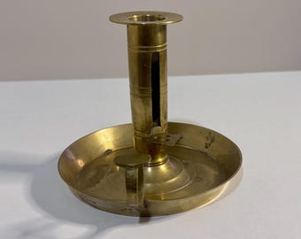 Könnte beinhalten: Ein Messing-Kerzenhalter mit breitem, kreisförmigem Tropfschalenfuß, einem zentralen zylindrischen Schaft mit vertikalem Schlitz, einem verschiebbaren Daumengriff zur Höhenverstellung der Kerze und einem kleinen, seitlich angebrachten Henkel.