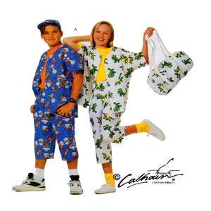 Könnte beinhalten: Zwei Kinder in passenden Pyjama-Sets. Eines ist blau mit orangefarbenen und gelben Figuren, das andere weiß mit grünen Froschgrafiken. Das Mädchen hält eine passende Tasche. Der Markenname "Calhoun Cotton Prints" ist sichtbar.