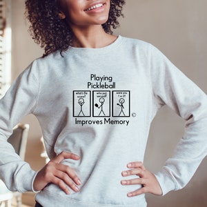 Puede incluir: Una sudadera gris con el texto "Playing Pickleball Improves Memory" y una ilustraci&oacute;n de un personaje de palo con cuatro paneles. Los paneles muestran personajes de palo con el texto "What's the score?", "Who just served?", "Were you 1 or 27?"