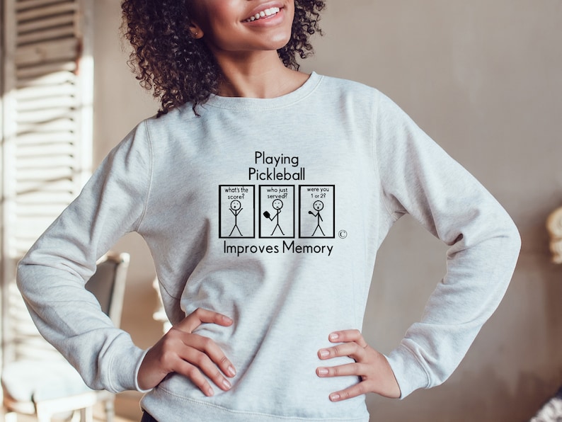 Puede incluir: Una sudadera gris con el texto "Playing Pickleball Improves Memory" y una ilustraci&oacute;n de un personaje de palo con cuatro paneles. Los paneles muestran personajes de palo con el texto "What's the score?", "Who just served?", "Were you 1 or 27?"