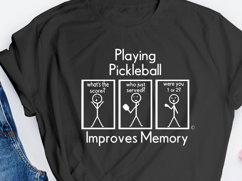 Puede incluir: Camiseta negra con texto blanco que dice "Jugar al pickleball mejora la memoria". El texto est&aacute; acompa&ntilde;ado por tres ilustraciones de figuras de palo.