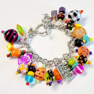 May include: A silver charm bracelet with a variety of Halloween-themed charms, including a pumpkin, a witch's hat, a candy corn, and a ghost. The charms are attached to a silver chain with a lobster clasp. The bracelet is decorated with colorful beads, including orange, green, purple, and black.