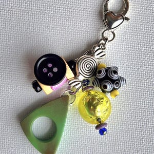 K&ouml;nnte beinhalten: Ein skurriler Charm mit einer Vielzahl dekorativer Elemente. Er enth&auml;lt einen schwarzen Knopf, eine silberne Spirale, ein gr&uuml;nes Dreieck und eine gelbe Perle. Der Charm ist an einer silbernen Kette mit einem herzf&ouml;rmigen Verschluss befestigt.