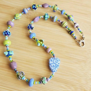 May include: A beaded necklace featuring a variety of glass beads in shades of blue, green, purple, and yellow. The beads include round, oval, and patterned shapes, with a prominent heart-shaped bead featuring blue swirls and a metal clasp.