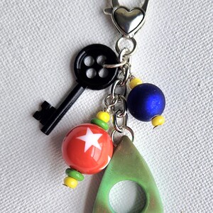 May include: A keychain with a silver heart-shaped clasp, featuring a black key, red and white star bead, blue bead, and a green triangle charm. Yellow and green beads accent the design.