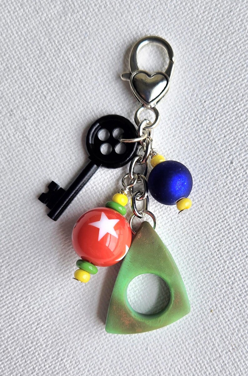 May include: A keychain with a silver heart-shaped clasp, featuring a black key, red and white star bead, blue bead, and a green triangle charm. Yellow and green beads accent the design.