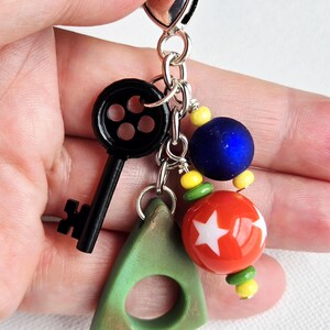May include: A silver keychain with a black key charm, a green triangle charm, and colorful beads. The beads include a blue sphere, a red sphere with white stars, and yellow and green beads. The keychain is held in a hand.