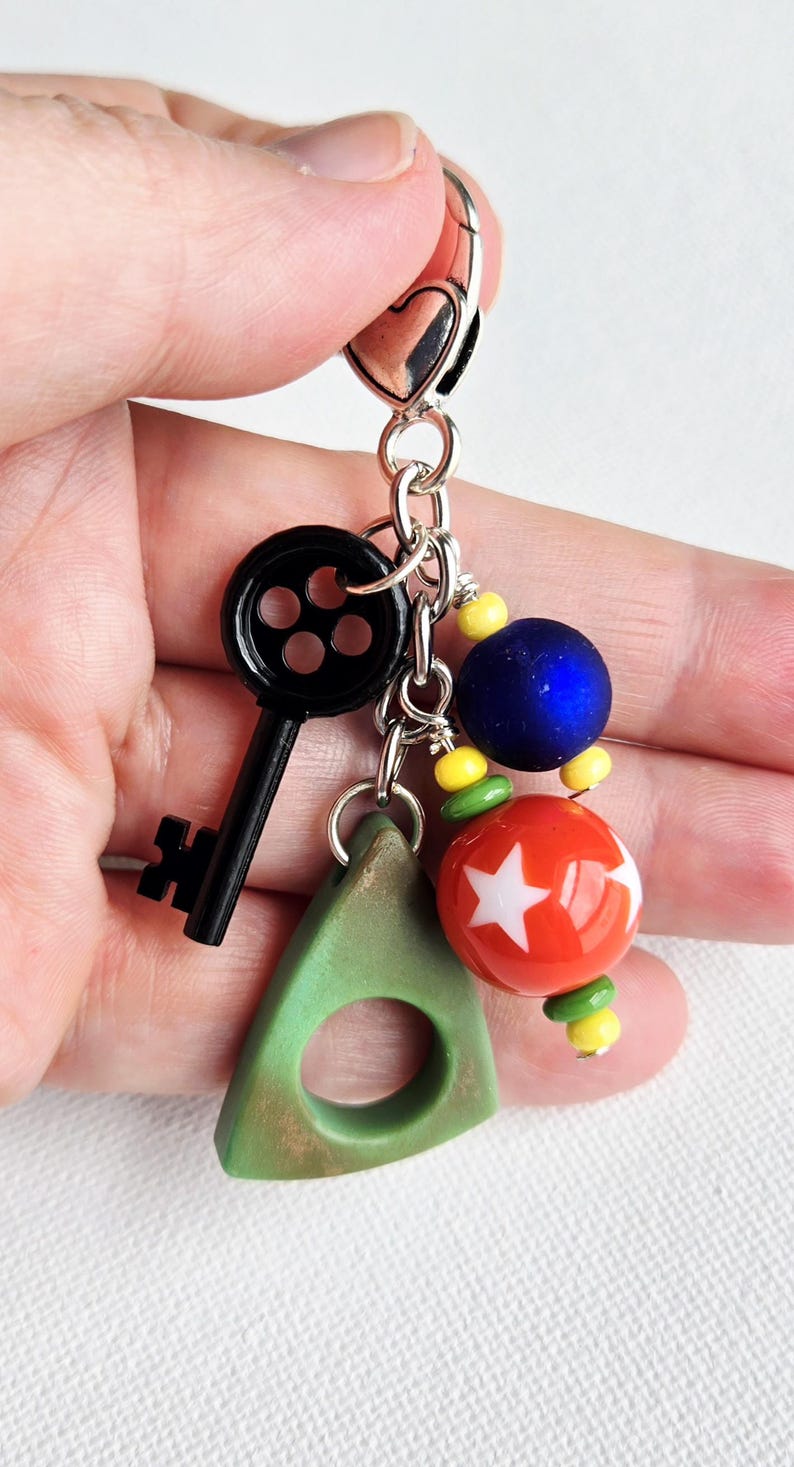 May include: A silver keychain with a black key charm, a green triangle charm, and colorful beads. The beads include a blue sphere, a red sphere with white stars, and yellow and green beads. The keychain is held in a hand.