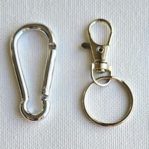 K&ouml;nnte beinhalten: Eine Auswahl an silberfarbenen Metallverschl&uuml;ssen und Zubeh&ouml;r. Dazu geh&ouml;ren ein kleiner Karabinerverschluss, ein Karabinerhaken, ein Drehverschluss mit Schl&uuml;sselring und ein schwarzes Schl&uuml;sselband mit kleinem Ring. Diese werden wahrscheinlich zum Befestigen von Gegenst&auml;nden verwendet.